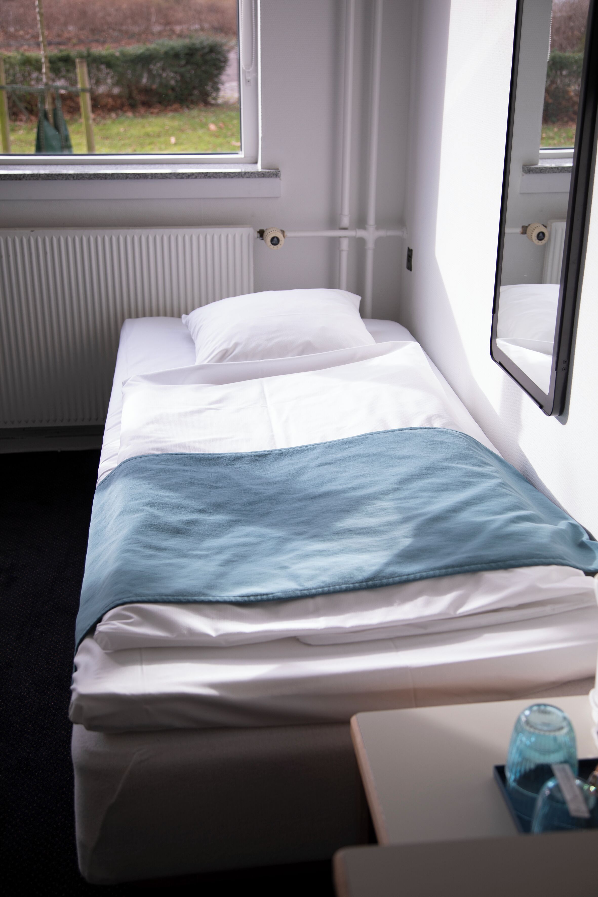A small, single-bed room with a window, a radiator, and a bedside table with a water bottle.