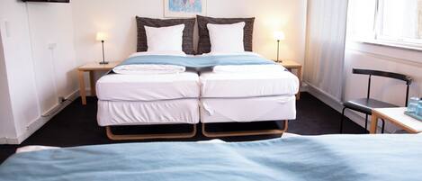 A double bed with a blue blanket, two bedside tables with lamps, a flat-screen TV on the wall, and a window with curtains.