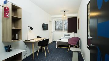 A compact hotel room with a bed, a desk with a computer, a chair, a bookshelf, and a window with a view of trees.