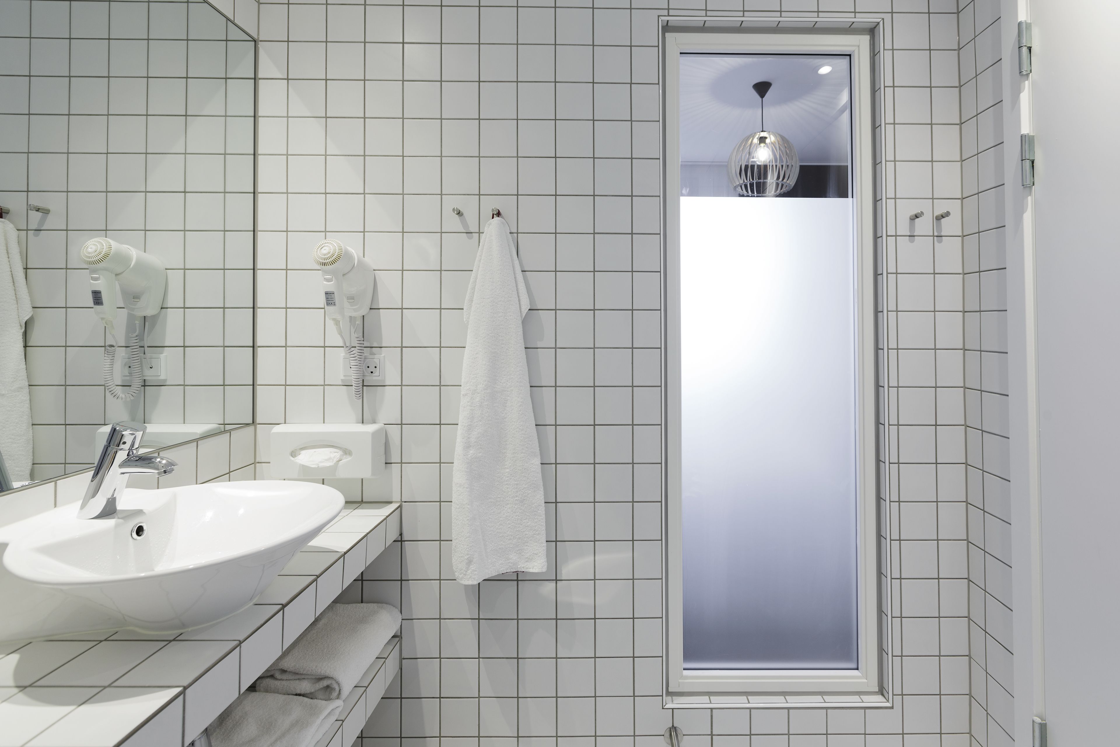 A modern bathroom with a white sink, two hair dryers, a towel rack, and a window with a view.