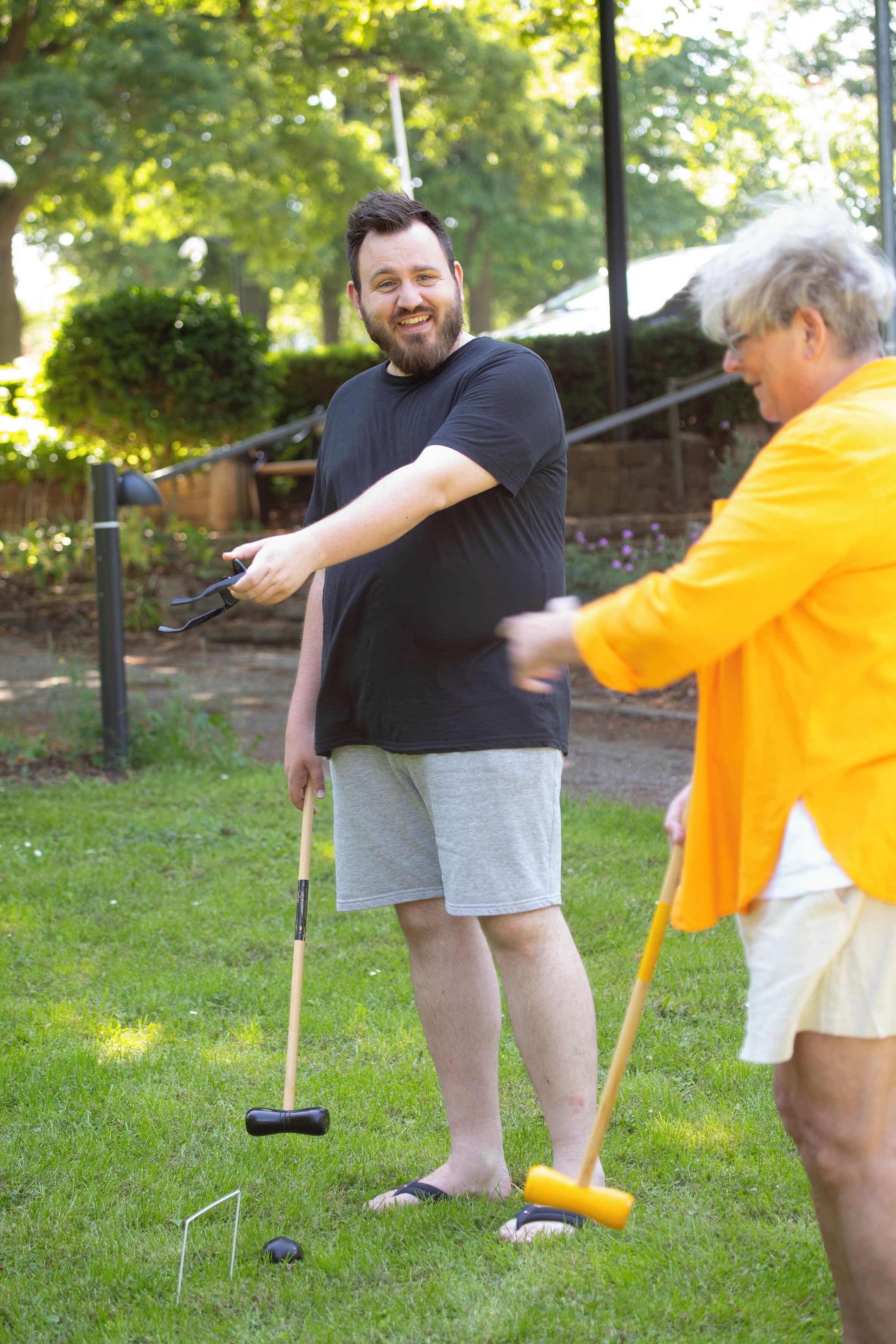 Two people playing croquet in a grassy outdoor area with trees and a fence in the background.
