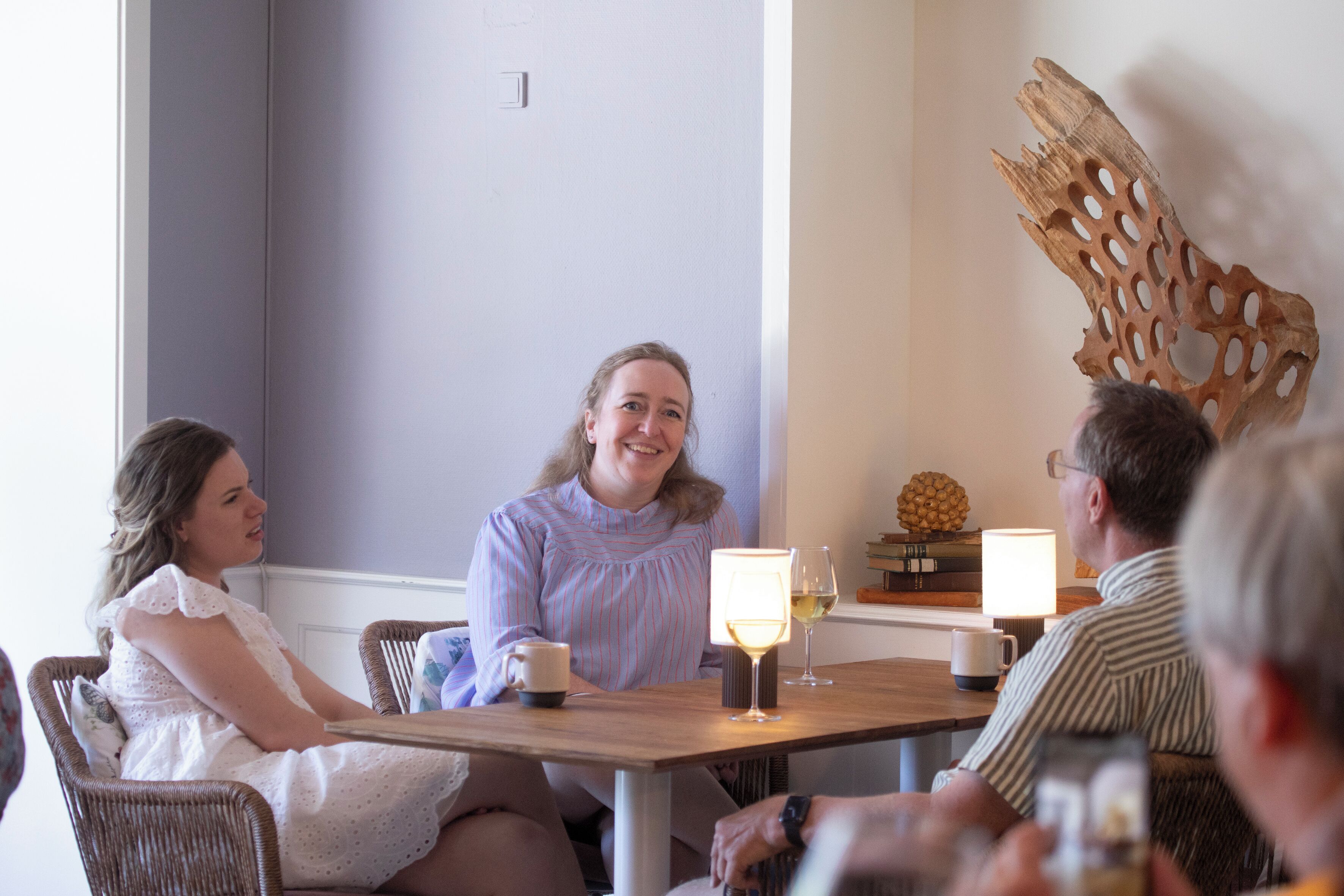 Un grupo de personas sentadas alrededor de una mesa de madera con dos lámparas encendidas, un adorno de madera en la pared y una planta pequeña sobre la mesa.