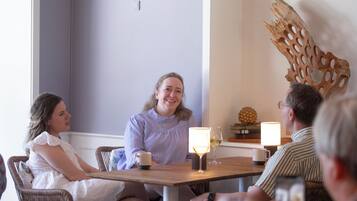 A group of people seated around a wooden table with two lit lamps, a decorative wooden piece on the wall, and a small plant on the table.