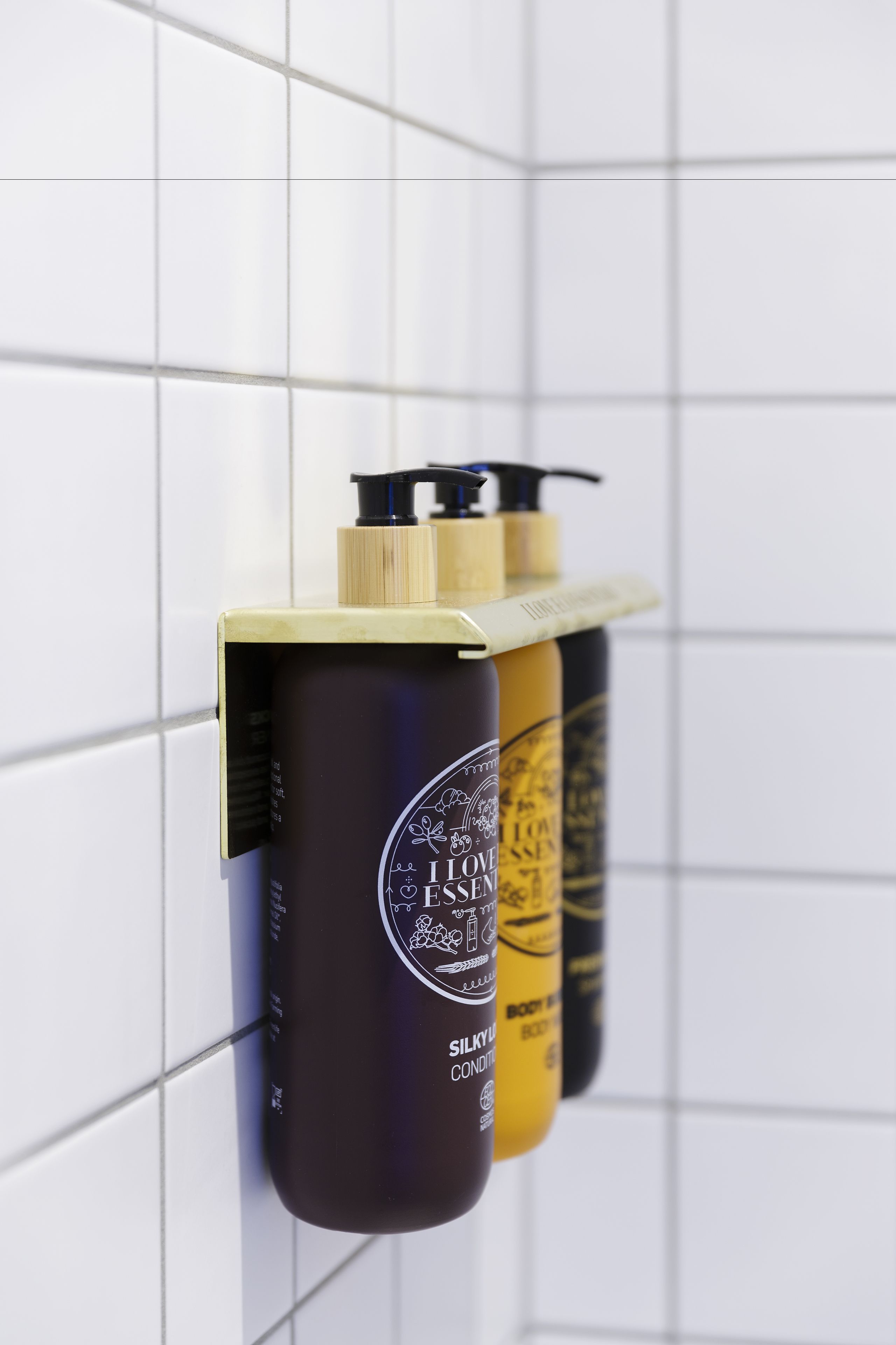 Drei Kosmetikflaschen mit Dosierern, beschriftet mit 'I Love Essence', hängen an einer gefliesten Wand.