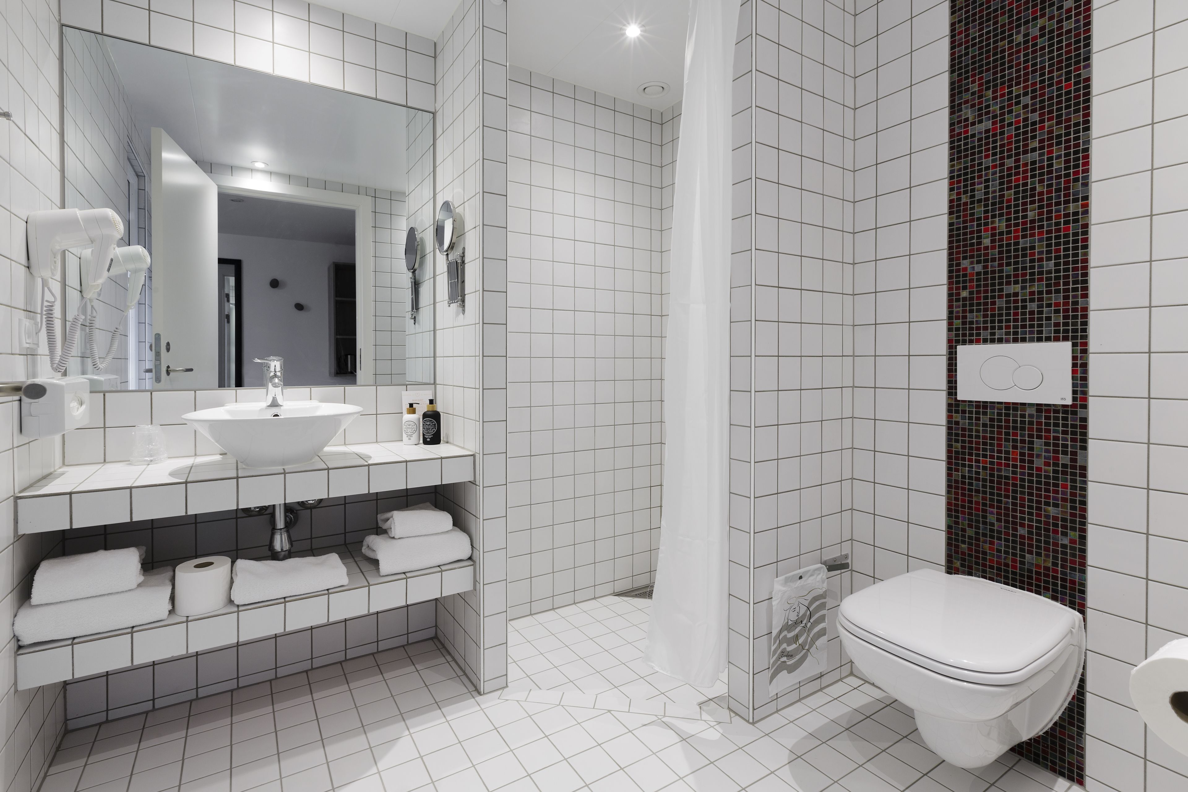 A modern bathroom with a white sink, mirror, and toilet. There is a shower area with a tiled wall and a curtain.