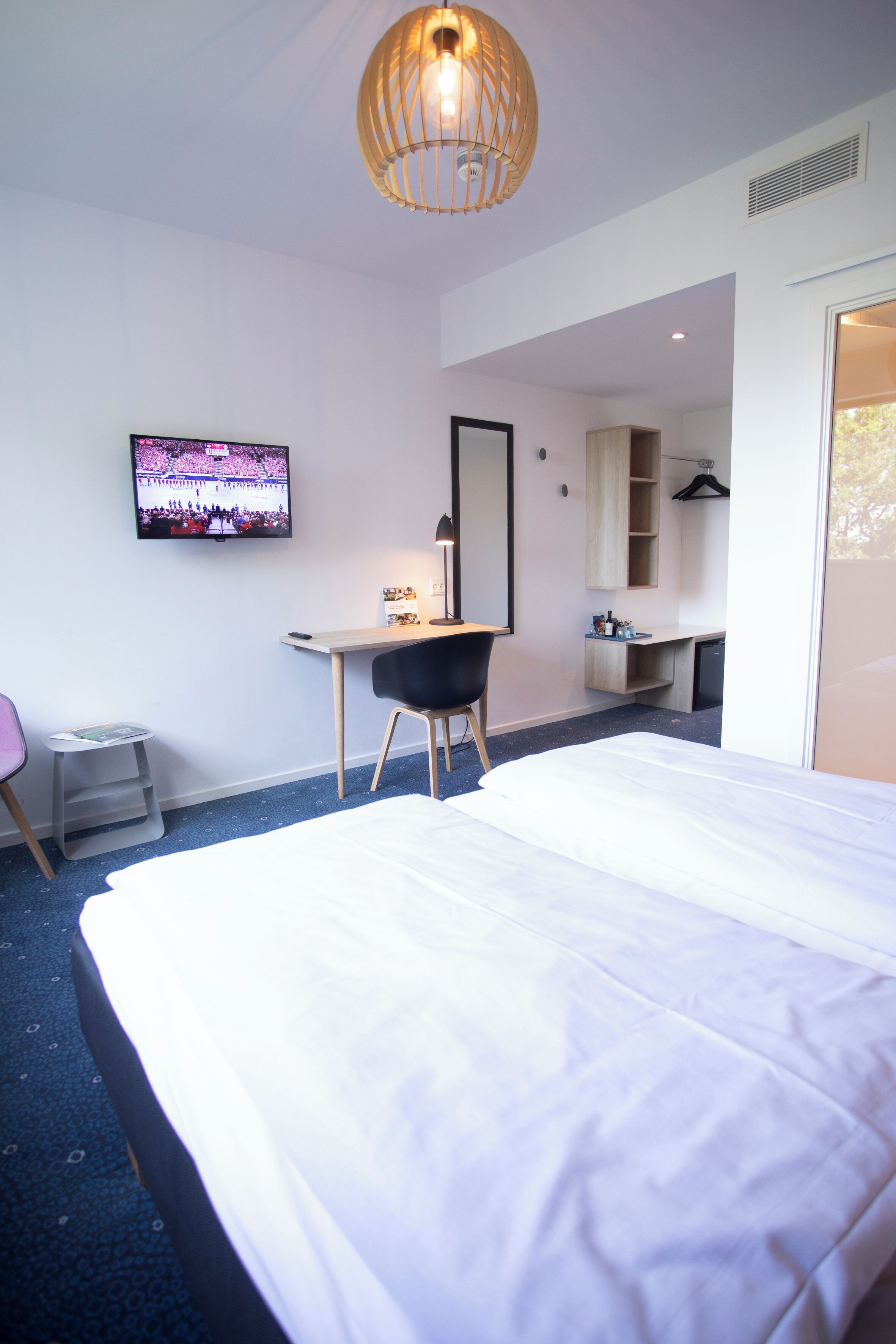 A modern hotel room with a large bed, a desk, a chair, a television, and a wardrobe.
