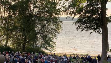 A large outdoor gathering of people by a lake, with trees and grass in the foreground.