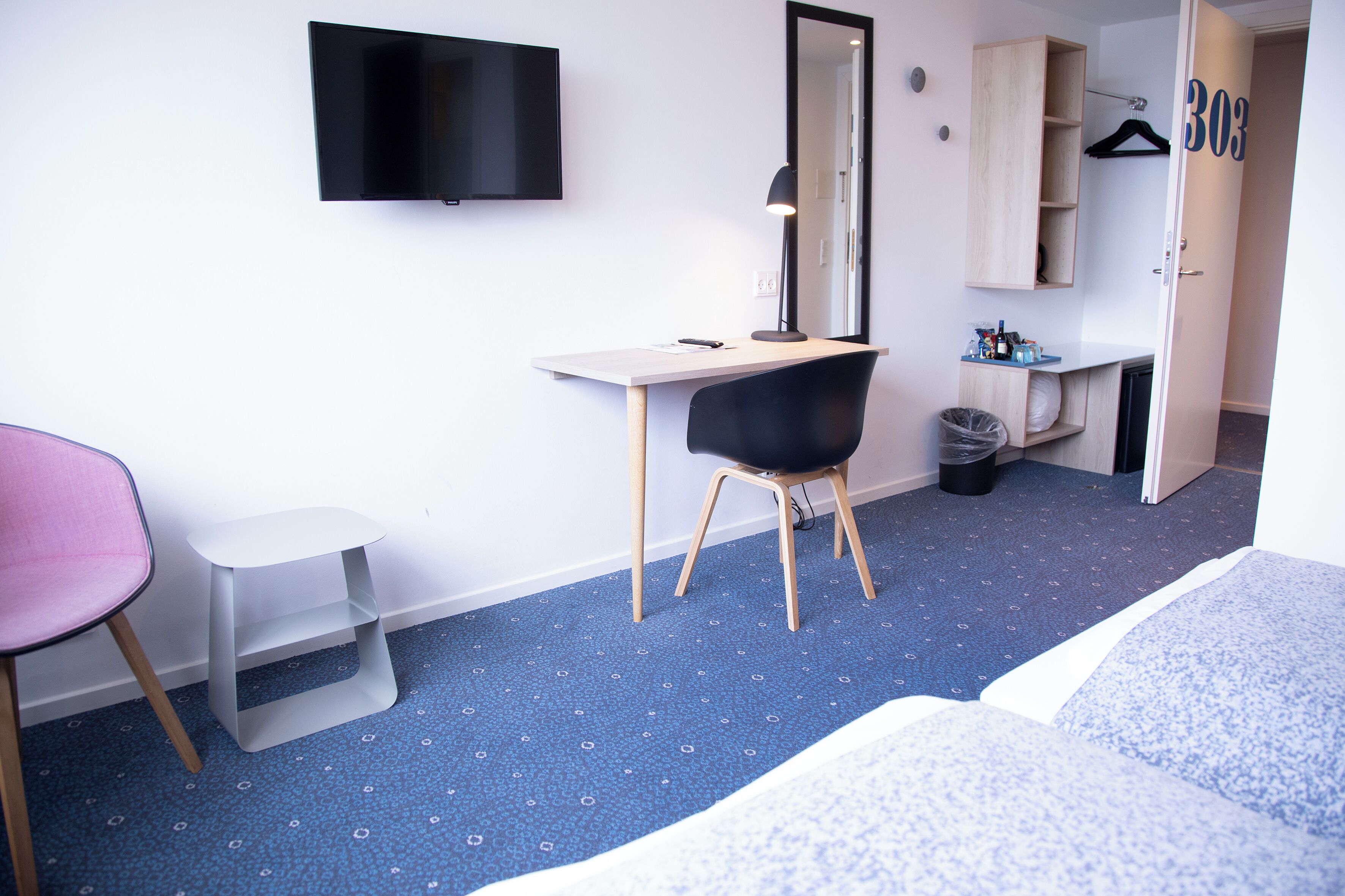 Una habitación de hotel con cama, escritorio y silla, televisor montado en la pared, espejo y armario.