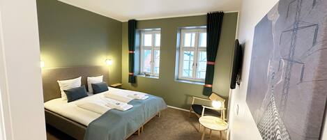 A compact hotel room with a bed, a desk, a chair, two windows with curtains, a wall-mounted TV, and a painting.