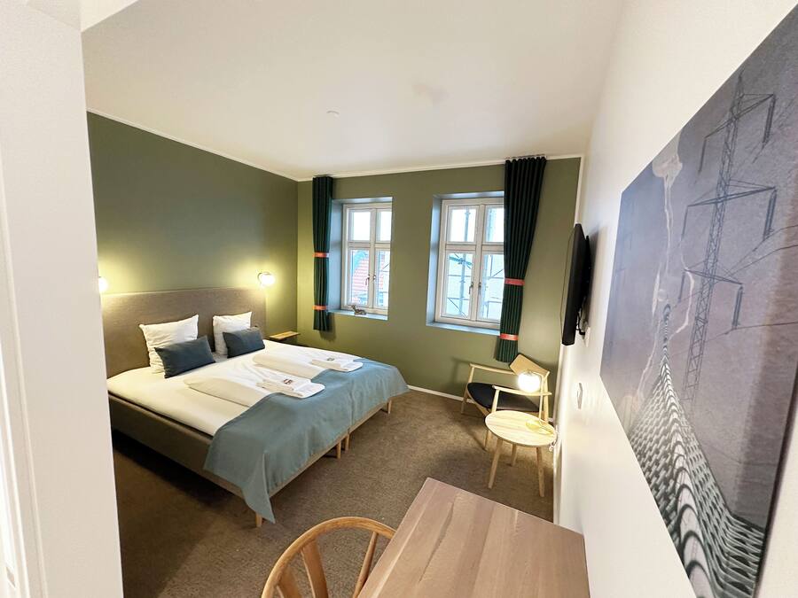 A compact hotel room with a bed, a desk, a chair, two windows with curtains, a wall-mounted TV, and a painting.