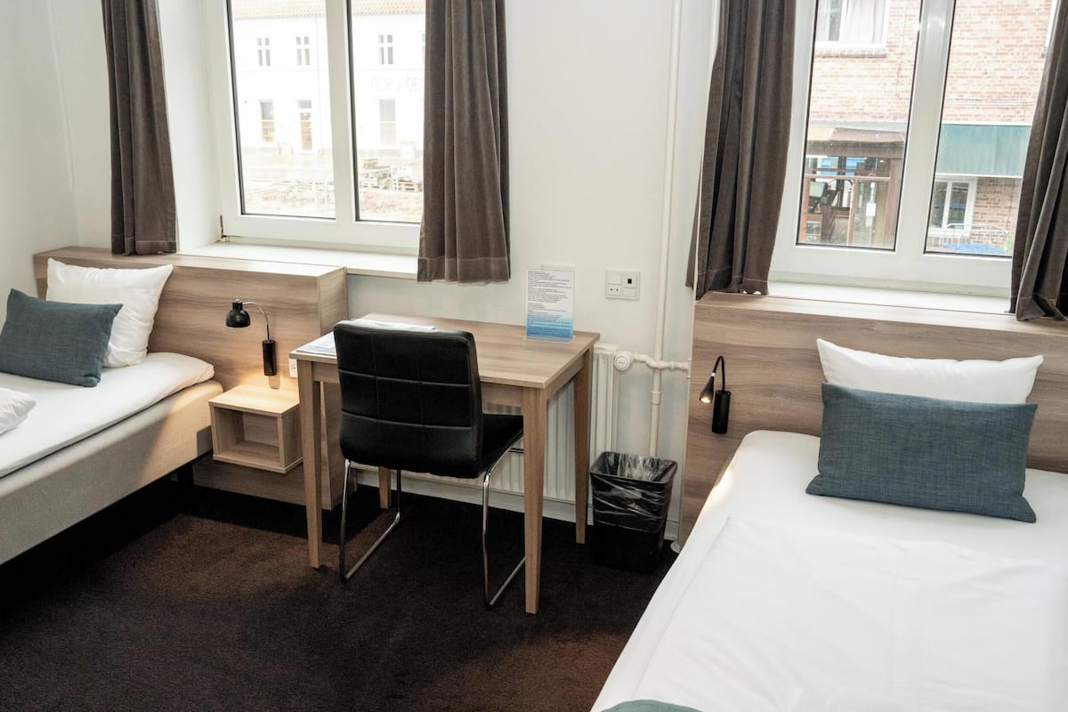 Ein Hotelzimmer mit zwei Einzelbetten, einem Schreibtisch, einem Stuhl und einer kleinen Mülleimer.