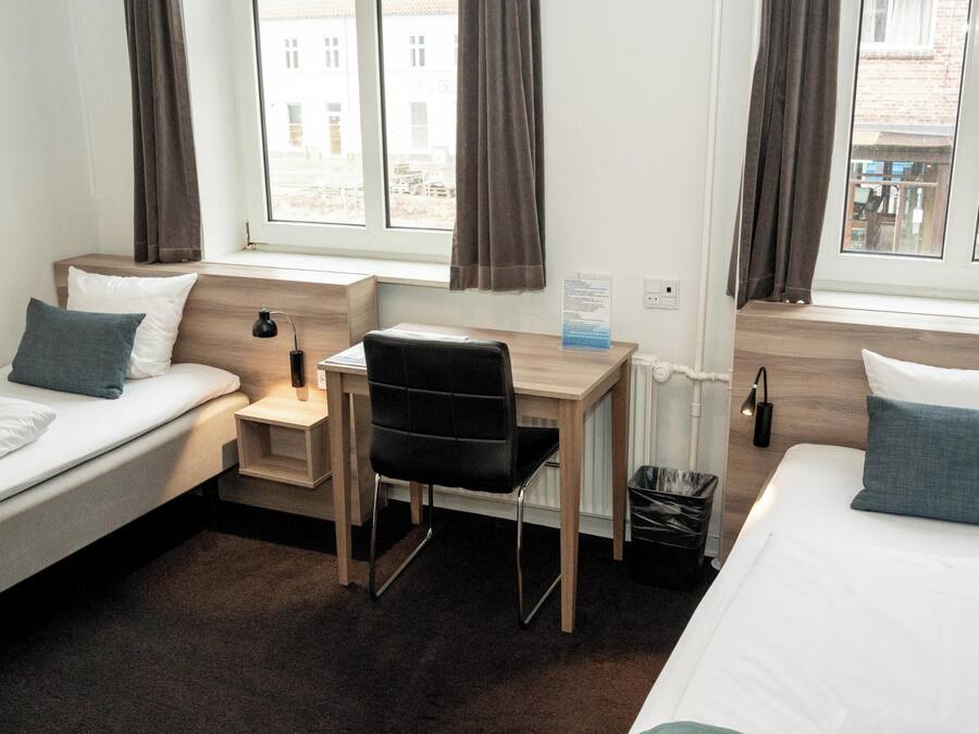 A hotel room with two single beds, a desk, a chair, and a small trash bin.