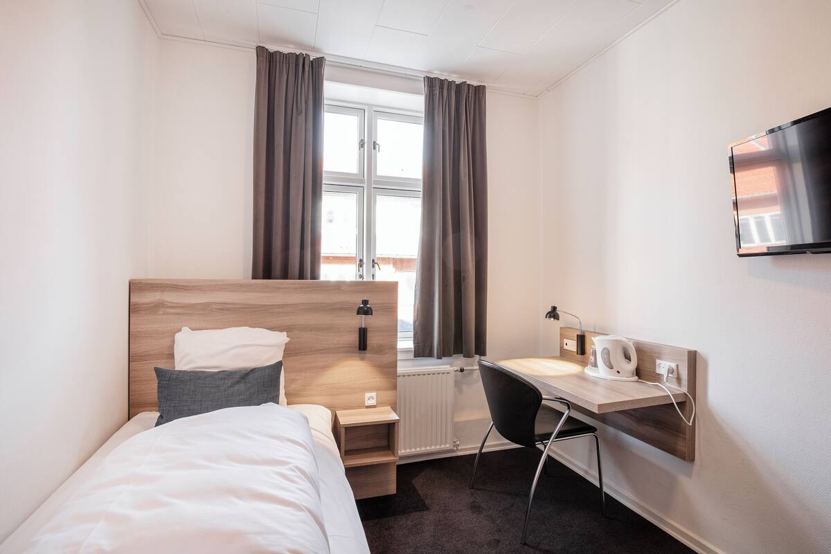Ein Hotelzimmer mit Bett, Schreibtisch, Stuhl, Fernseher und einem Fenster mit Vorhängen.