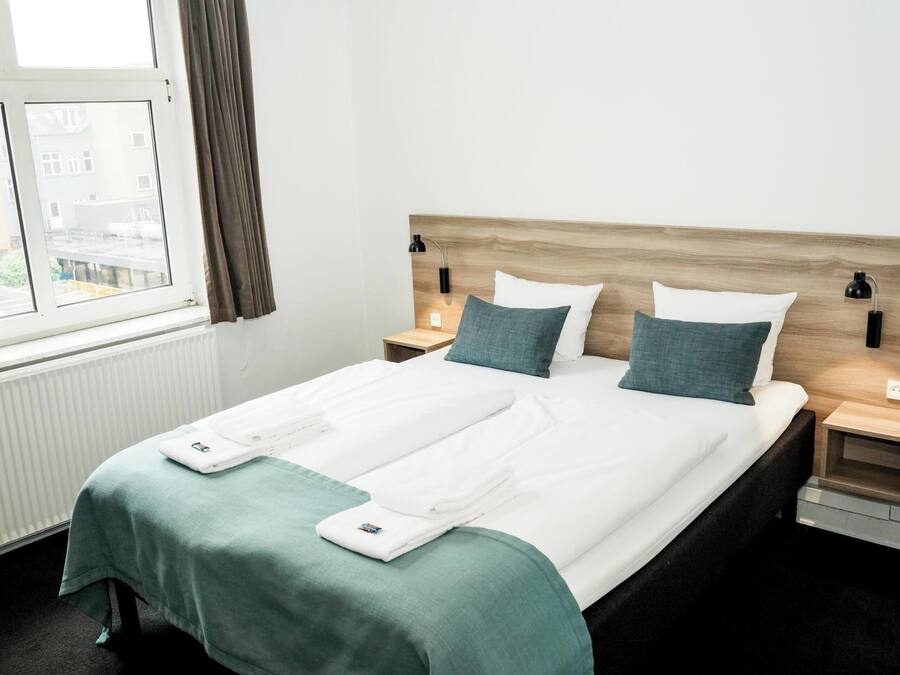 A hotel room with a large bed, two bedside tables, a window with curtains, and a view of buildings outside.
