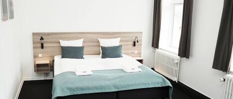 A neatly made bed with a teal bedspread and two pillows, a bedside table with a lamp, and a window with curtains.