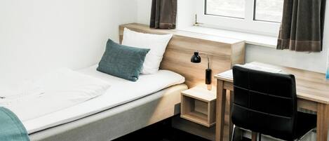 A bedroom with a bed, a desk, and a chair. There is a window with curtains, a nightstand with a lamp, and a view of buildings outside.