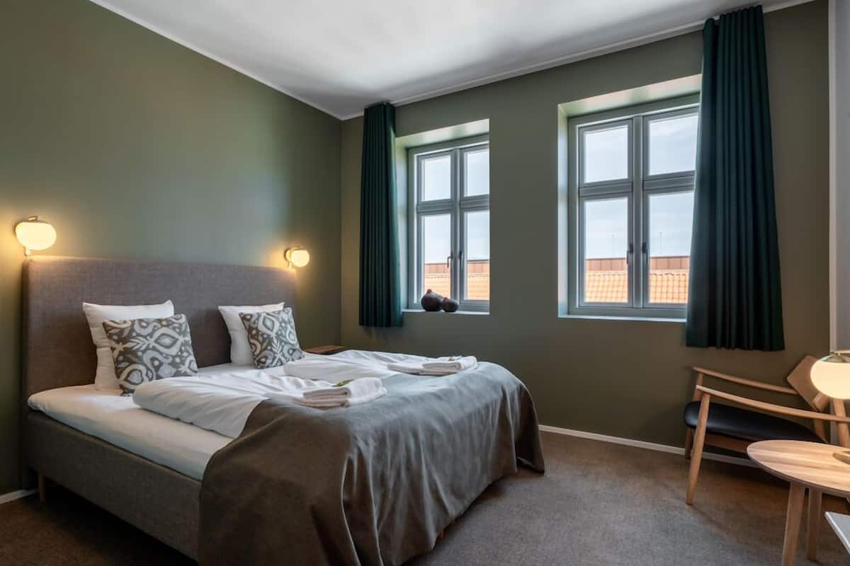 Ein Schlafzimmer mit einem Bett, zwei Nachttischlampen, einem Sessel, einem Tisch und zwei mit Vorhängen versehenen Fenstern.