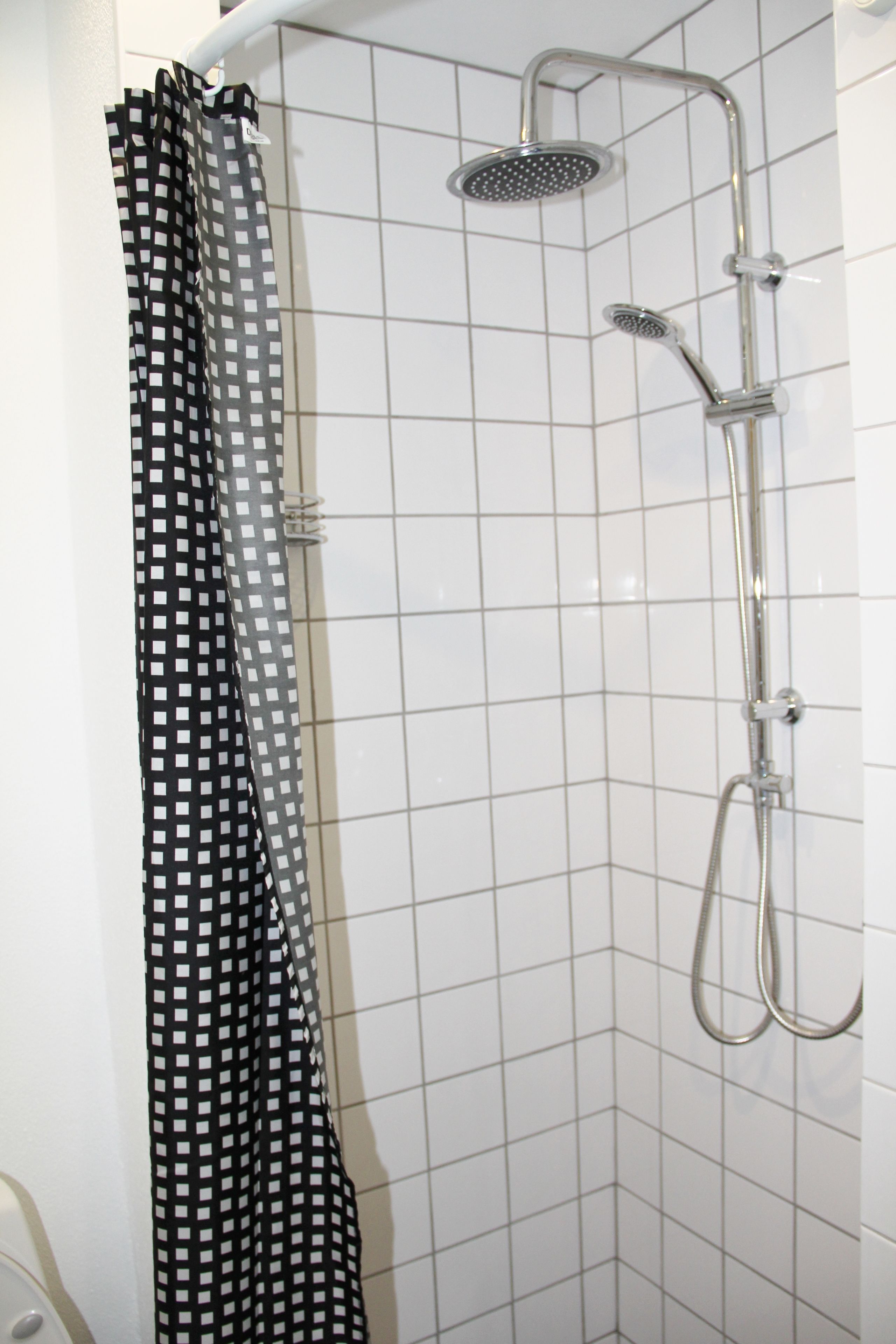 A shower with a handheld showerhead and a wall-mounted head, a checkered shower curtain, and white tiled walls.