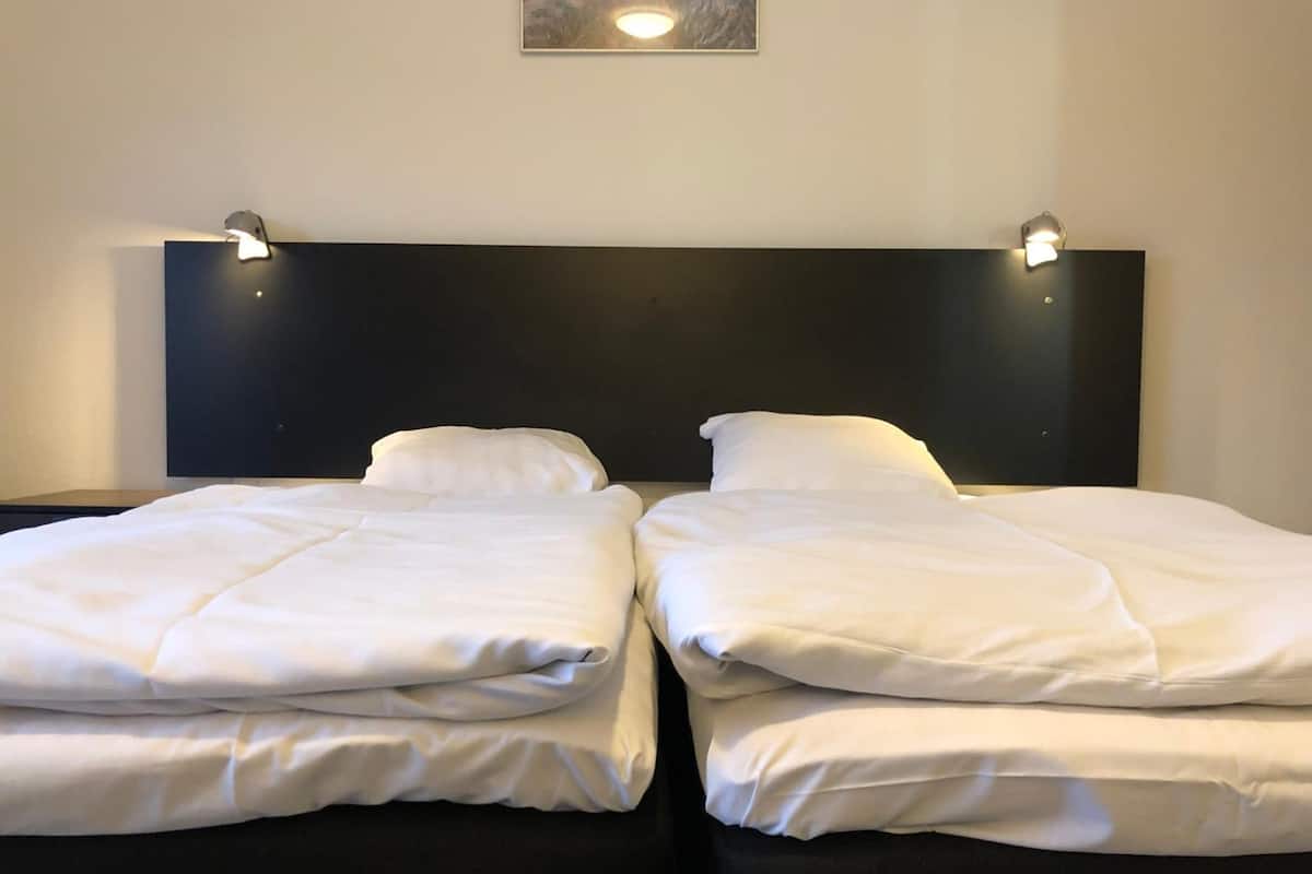 Una cama doble con sábanas blancas, cabecera oscura y dos lámparas de lectura fijadas a la pared.