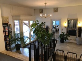 Un interior amplio con una escalera, varias plantas en macetas, una araña y un comedor visible a través de puertas de cristal.