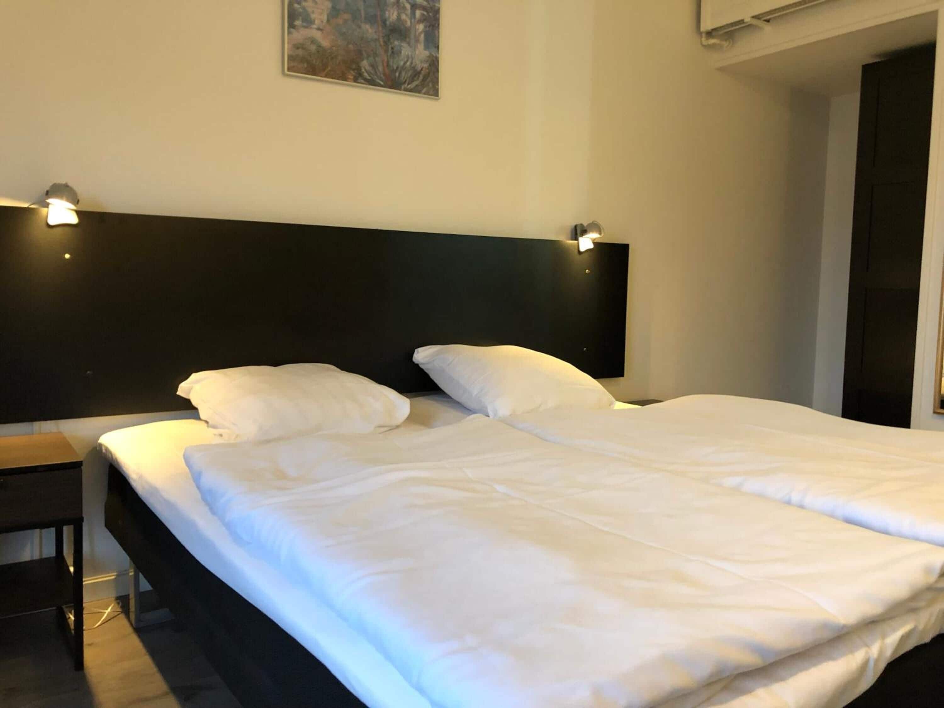 Una habitación de hotel con una cama grande, dos mesitas de noche, un cabecero con luces empotradas y un cuadro en la pared.