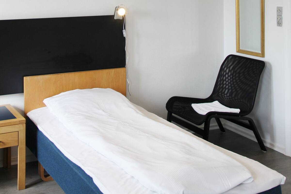 Una cama individual con cabecera de madera, una silla negra, una mesita de noche y un espejo instalado en la pared.