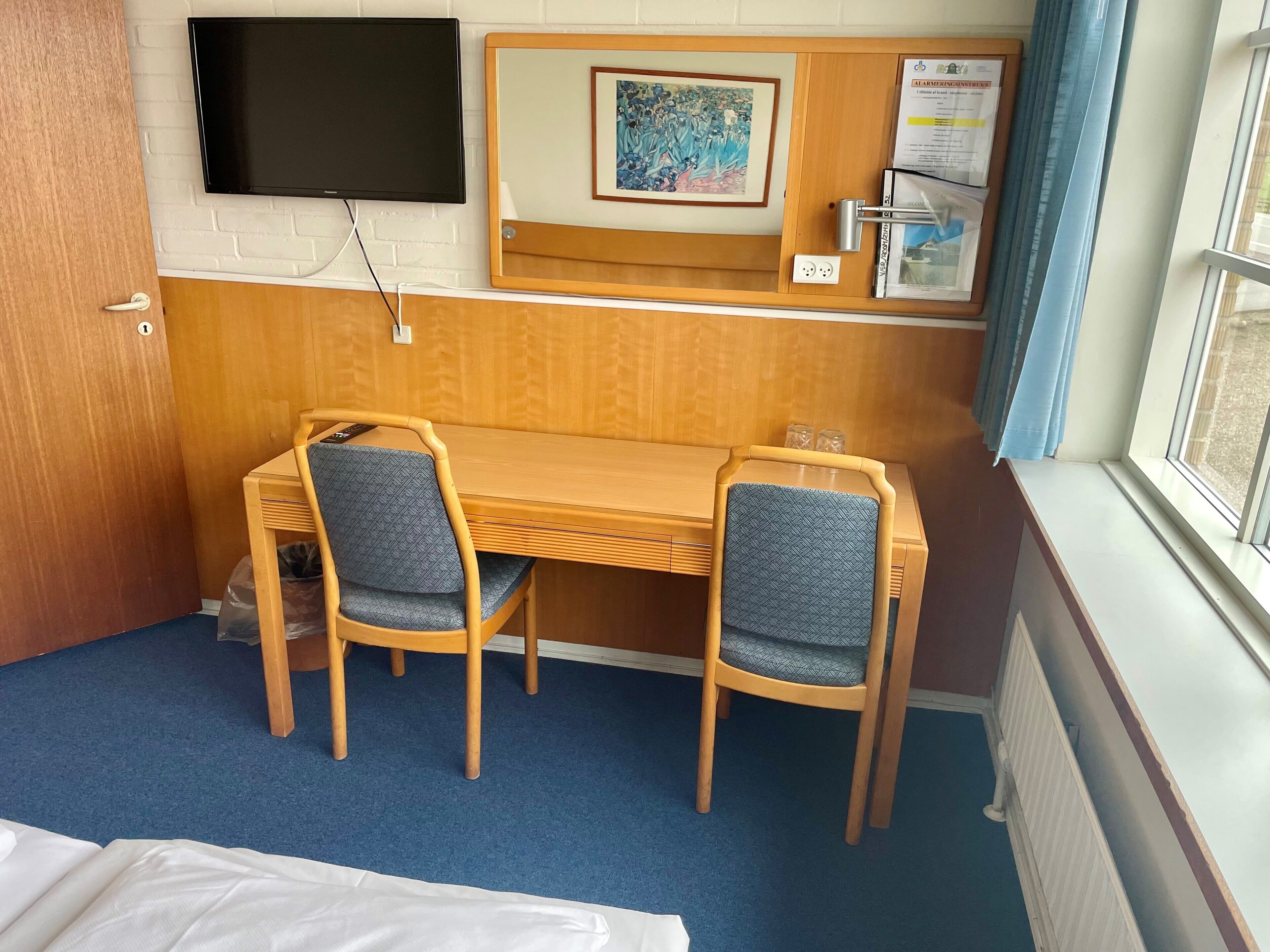 A hotel room with a wooden desk, two chairs, a TV mounted on the wall, a framed artwork, and a window with curtains.