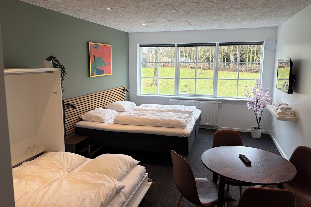 A hotel room with two beds, a small table, chairs, a window with a view of a grassy area, and a painting on the wall.