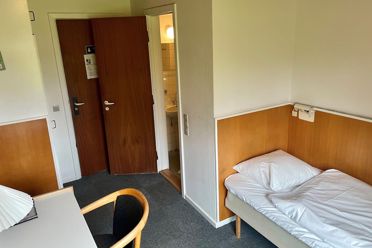A small, compact hotel room with a single bed, a wooden headboard, a small table, a chair, and a door leading to a bathroom.