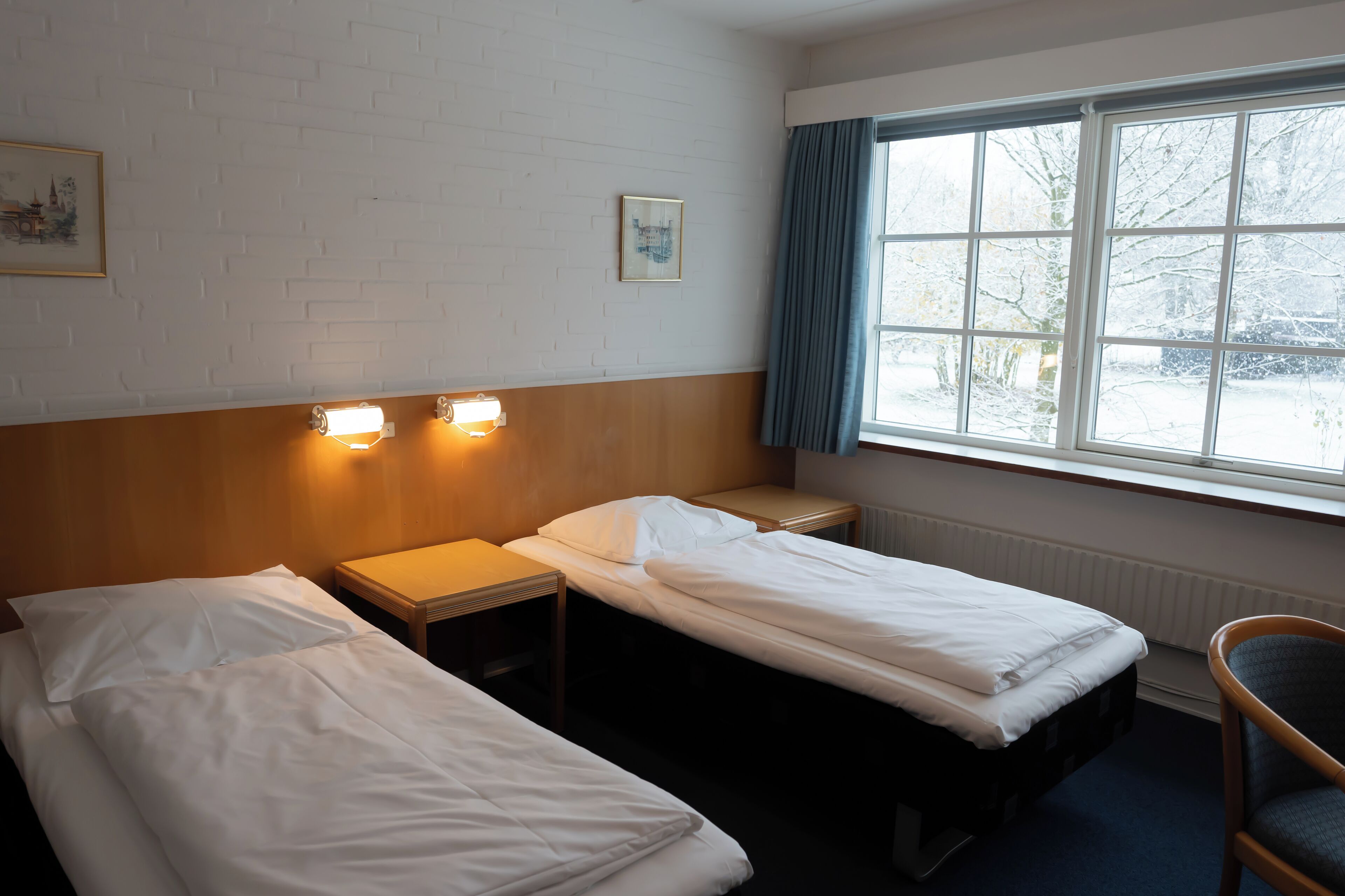 Et hotelværelse med to senge, en træsenggavl, et vindue med udsigt til et snelandskab og en stol.