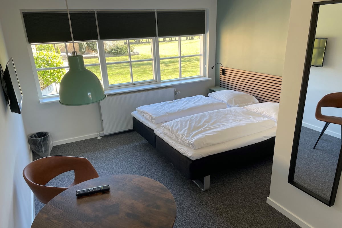 A bedroom with two beds, a window with blinds, a radiator, a wooden table, a chair, and a mirror.