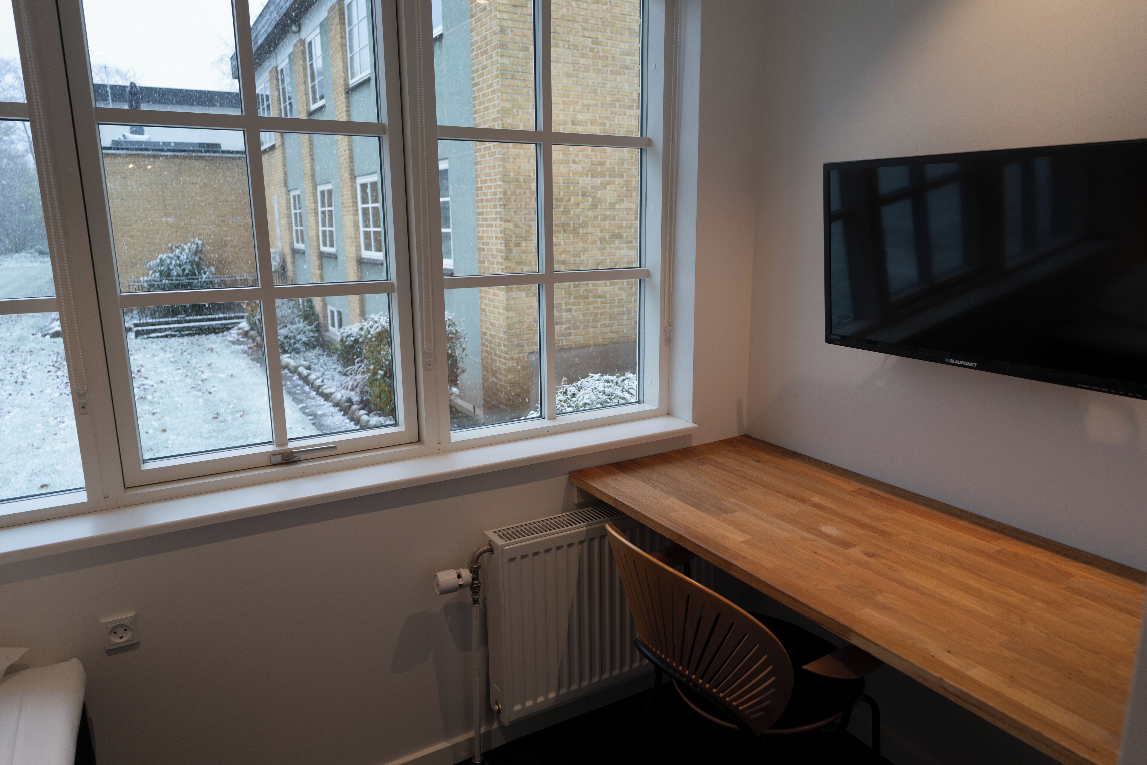Una habitación con un ventanal grande, un escritorio de madera, una silla y un televisor de pantalla plana montado.
