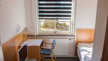 A small, compact room with a wooden desk, a blue chair, a bed, and a window with blinds.