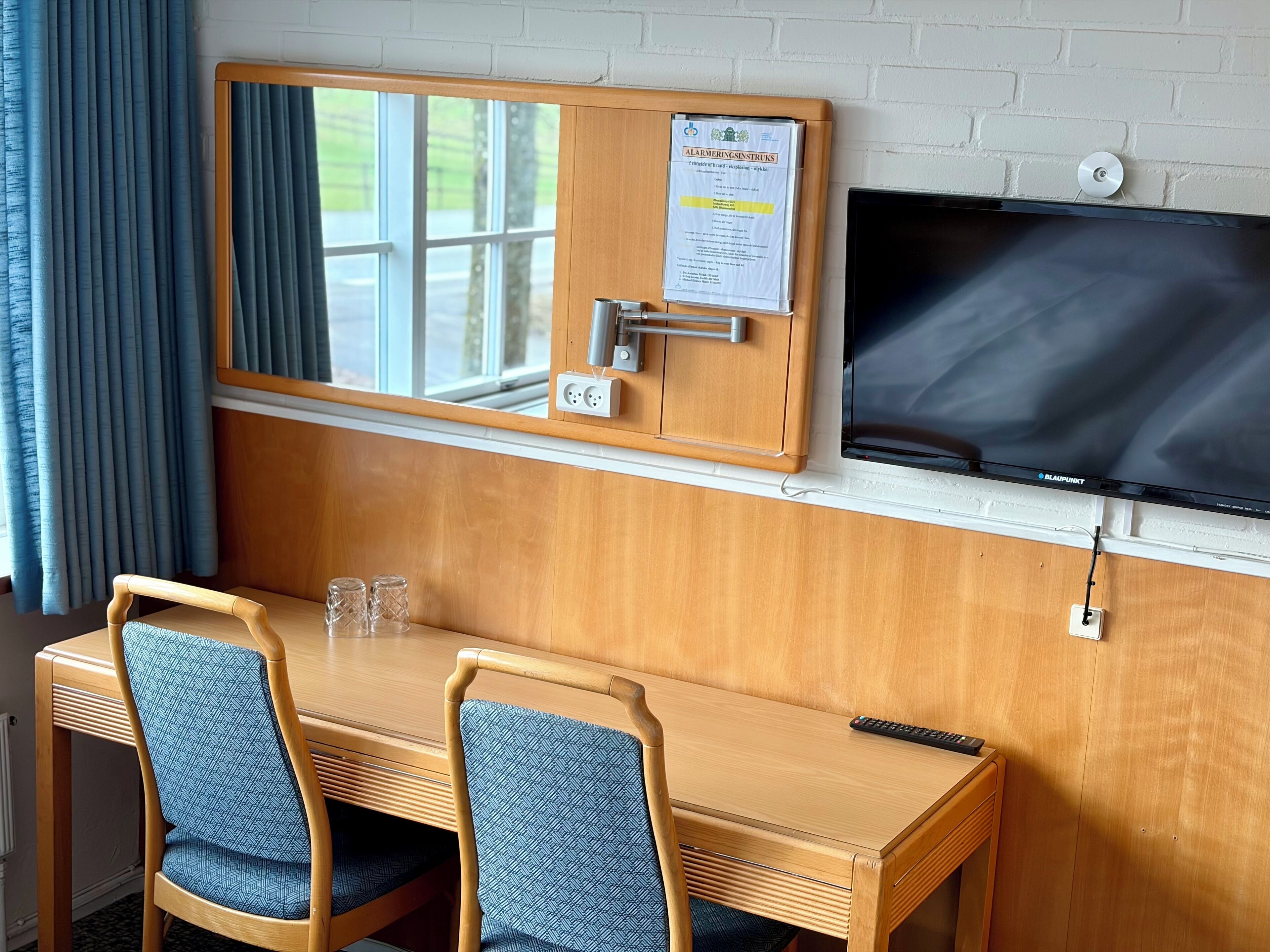 A hotel room with a wooden table, two chairs, a TV mounted on the wall, a window with curtains, and a wall-mounted alarm clock.