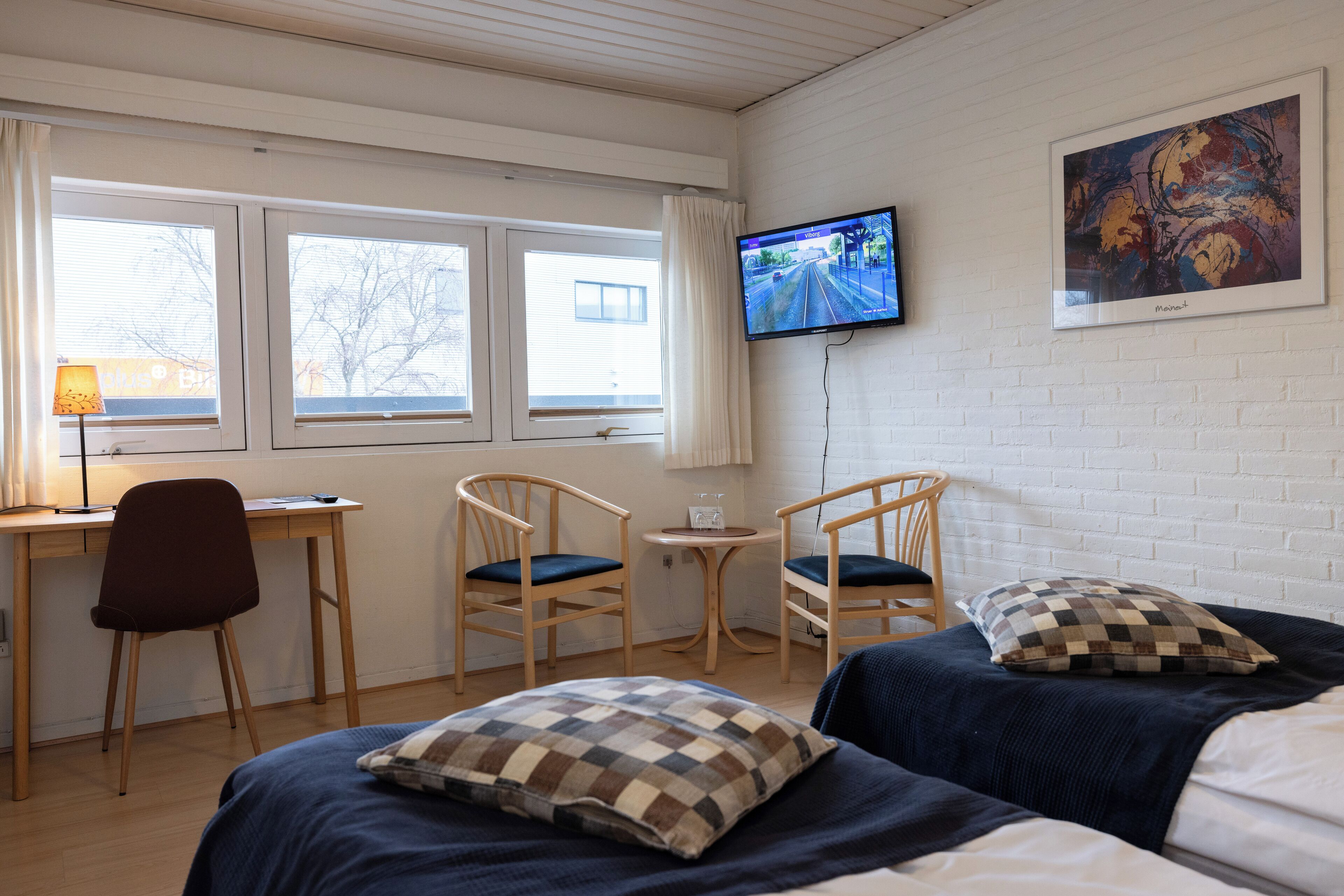 A hotel room with two beds, a desk, two chairs, a TV, and a window with curtains.