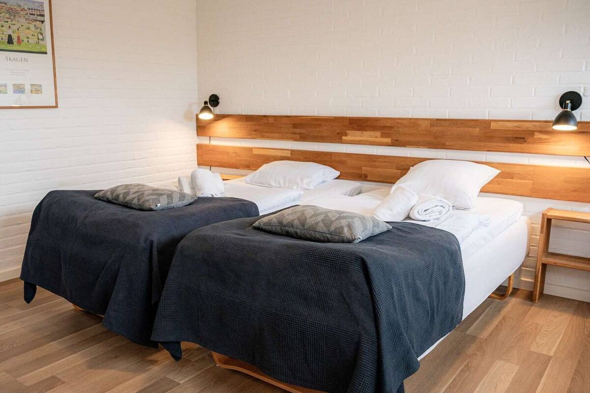 Une chambre double avec deux lits, chacun doté d’une literie et de coussins aux couleurs sombres, d’une tête de lit en bois et d’un mur en briques blanches.