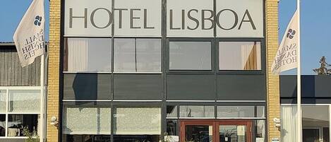 Ein Hotel namens „HOTEL LISBOA“ mit einer modernen Fassade, großen Fenstern und einem gut sichtbaren Eingang.