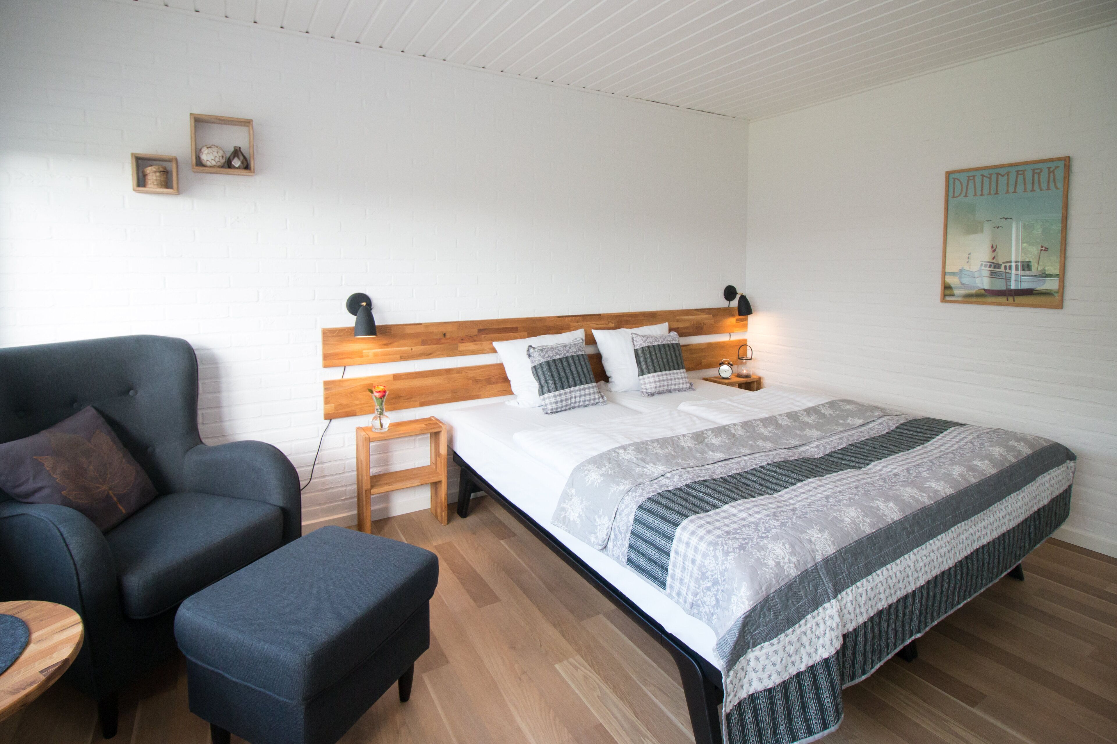 Un dormitorio con cabecera de madera, cama con colcha a rayas, un sillón oscuro, una mesita de madera y una mesita de noche con lámpara.
