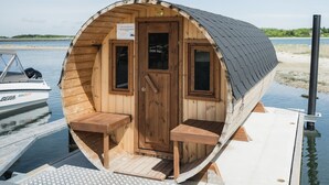 Eine hölzerne Hütte mit Tür und zwei Fenstern, einer Bank und einem kleinen Tisch auf einem Steg am Wasser.