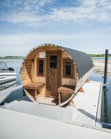 A wooden cabin with a door and two windows, a bench, and a small table on a dock by water.