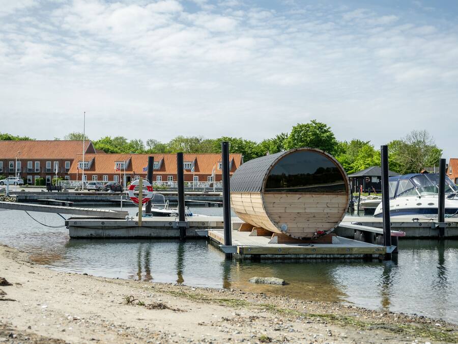 A waterfront dock with a unique wooden barrel structure, a marina with boats, and red-roofed buildings in the background.