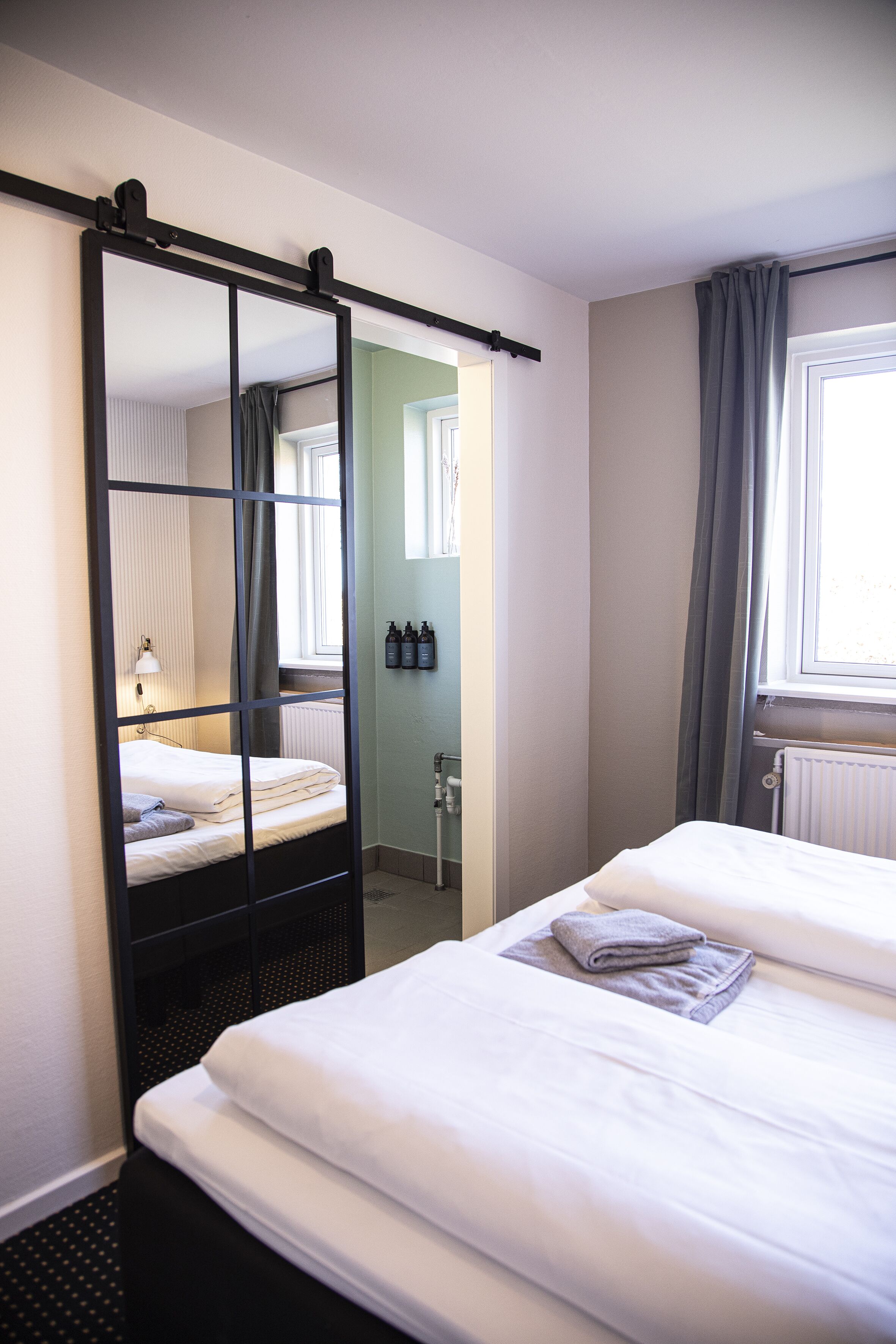 A hotel room with two beds, a large mirror, a window with curtains, and a radiator.