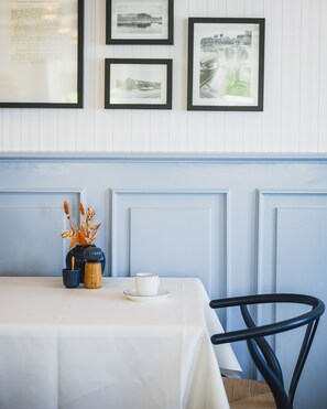 Un coin repas avec une table dressée pour deux personnes, un vase contenant des fleurs séchées et des tableaux encadrés accrochés au mur.
