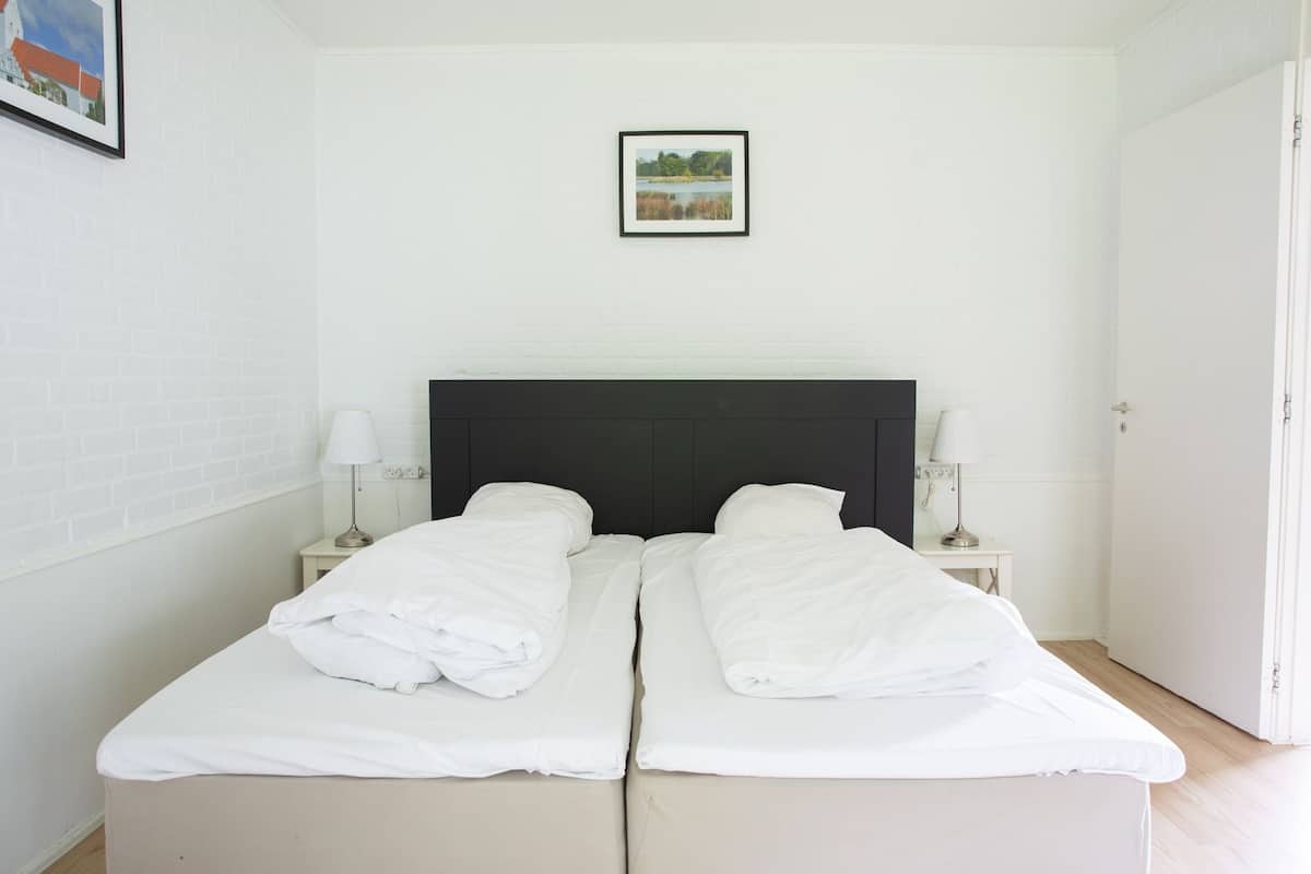 Una cama doble con ropa blanca, dos mesitas de noche con lámparas, un cuadro en la pared y una puerta a la derecha.