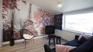 A modern living room with a large mural of the Eiffel Tower, a black TV stand, a black sofa, and a window with a view of the city. - Hotel Hedegaarden (Vejle)