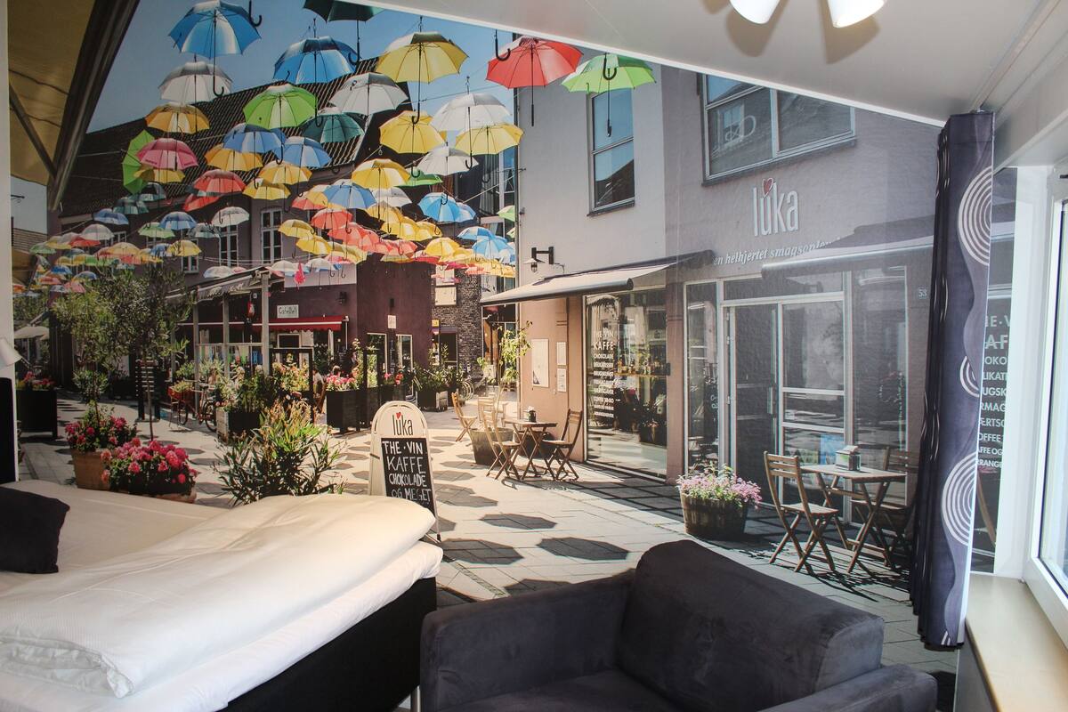 A hotel lobby with a large mural of colorful umbrellas, a seating area with tables and chairs, and a visible storefront with the name 'Luka'.