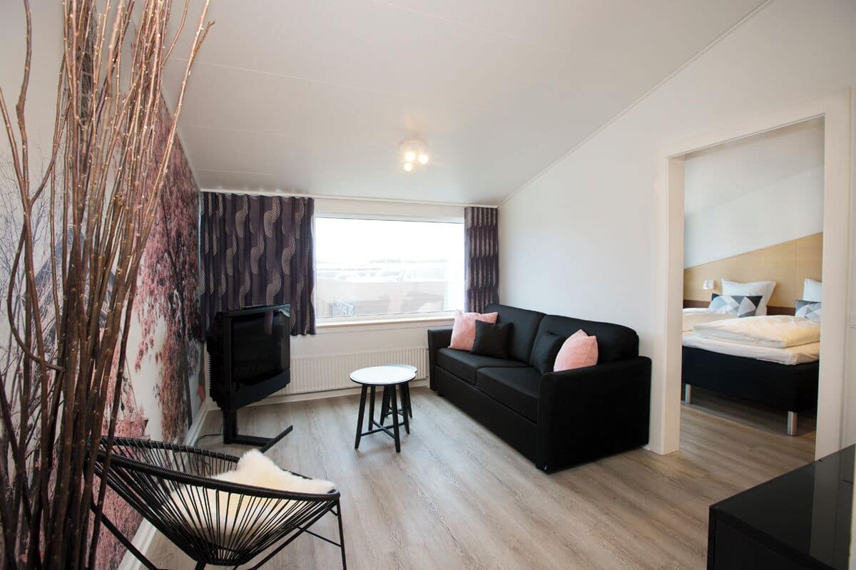 A modern hotel room with a black sofa, a small round table, a TV, and a bed with a wooden headboard.