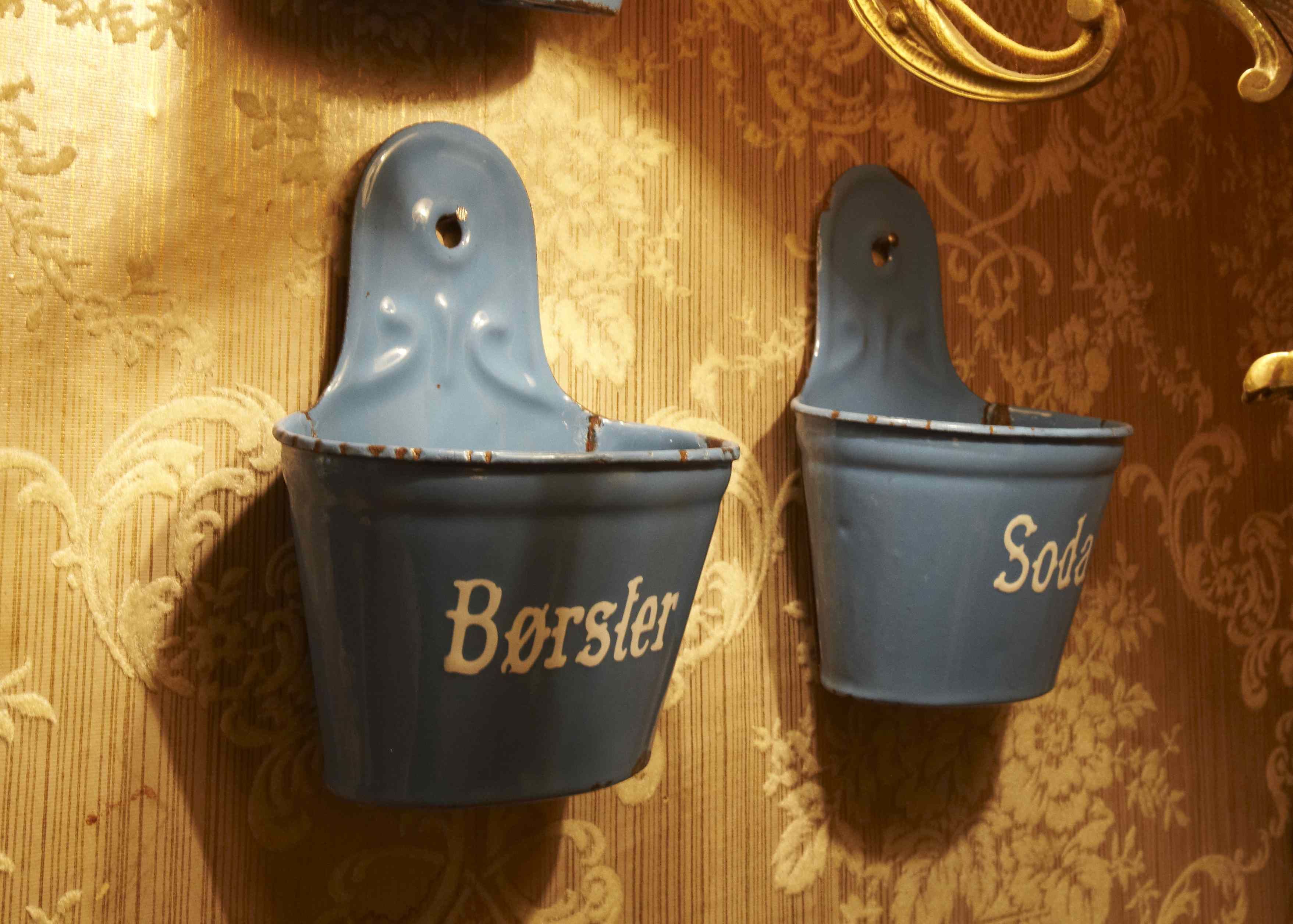 Two blue metal buckets with handles, one labeled "Borscht" and the other "Soda," mounted on a wall with a patterned wallpaper background.
