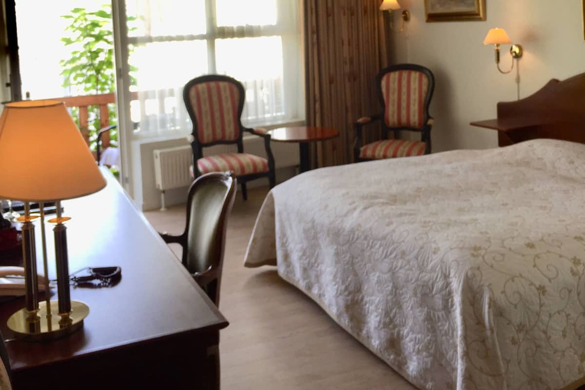 Une chambre d’hôtel avec un grand lit, un bureau avec une lampe, deux chaises, une petite table et une fenêtre avec des rideaux.