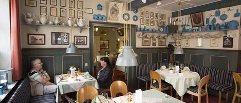 L’intérieur d’un restaurant présente des bancs à rayures, des tables recouvertes de nappes blanches et des étagères ornées de vaisselle en céramique bleue et blanche.